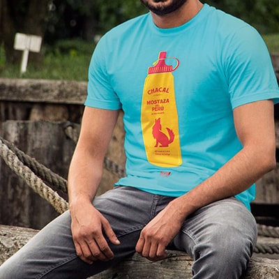 Fotografía en color, en primer plano, de una camiseta azul con el diseño Mostacero original de Moscas de Colores, que lleva un chico sentado en una valla.