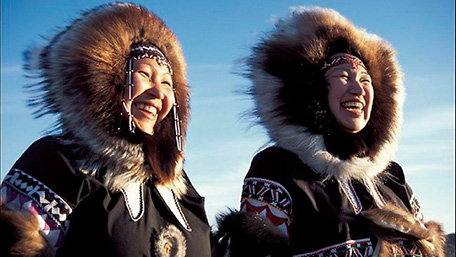 Two smiling Inuit women, dressed in their typical coats, under blue sky.