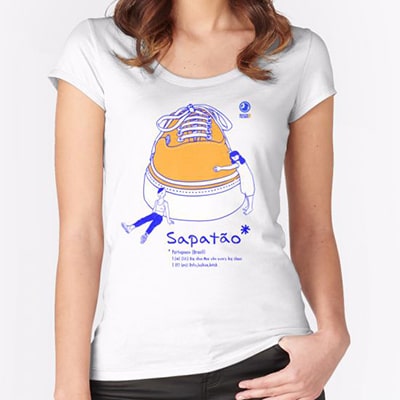 Close-up photograph of a white T-shirt worn by a girl with the Sapatão drawing, a big sneaker with two small girls around, with slang in large letters on the bottom. Drawn in blue and orange colors.